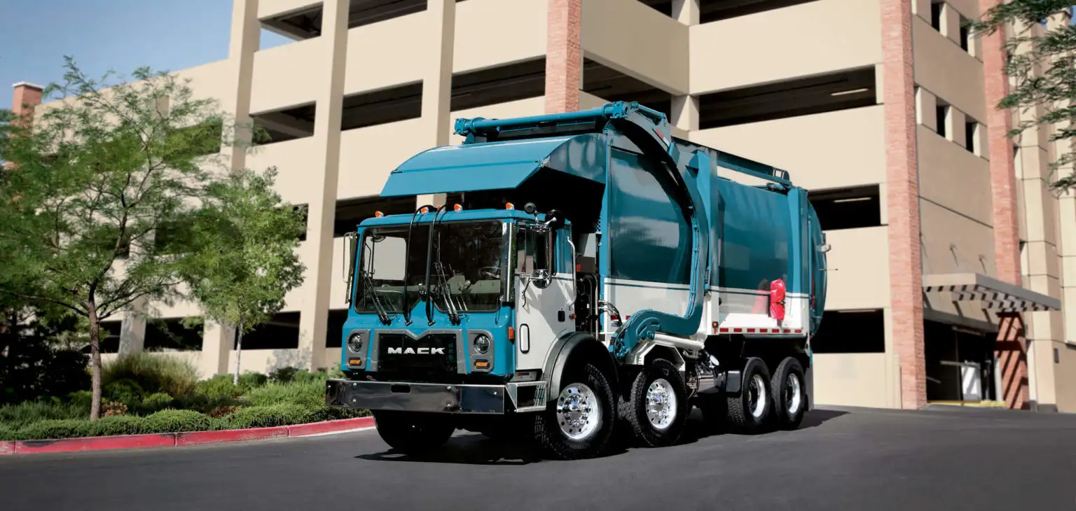Turquoise Mack Terrapro refuse truck driving past a parking structure