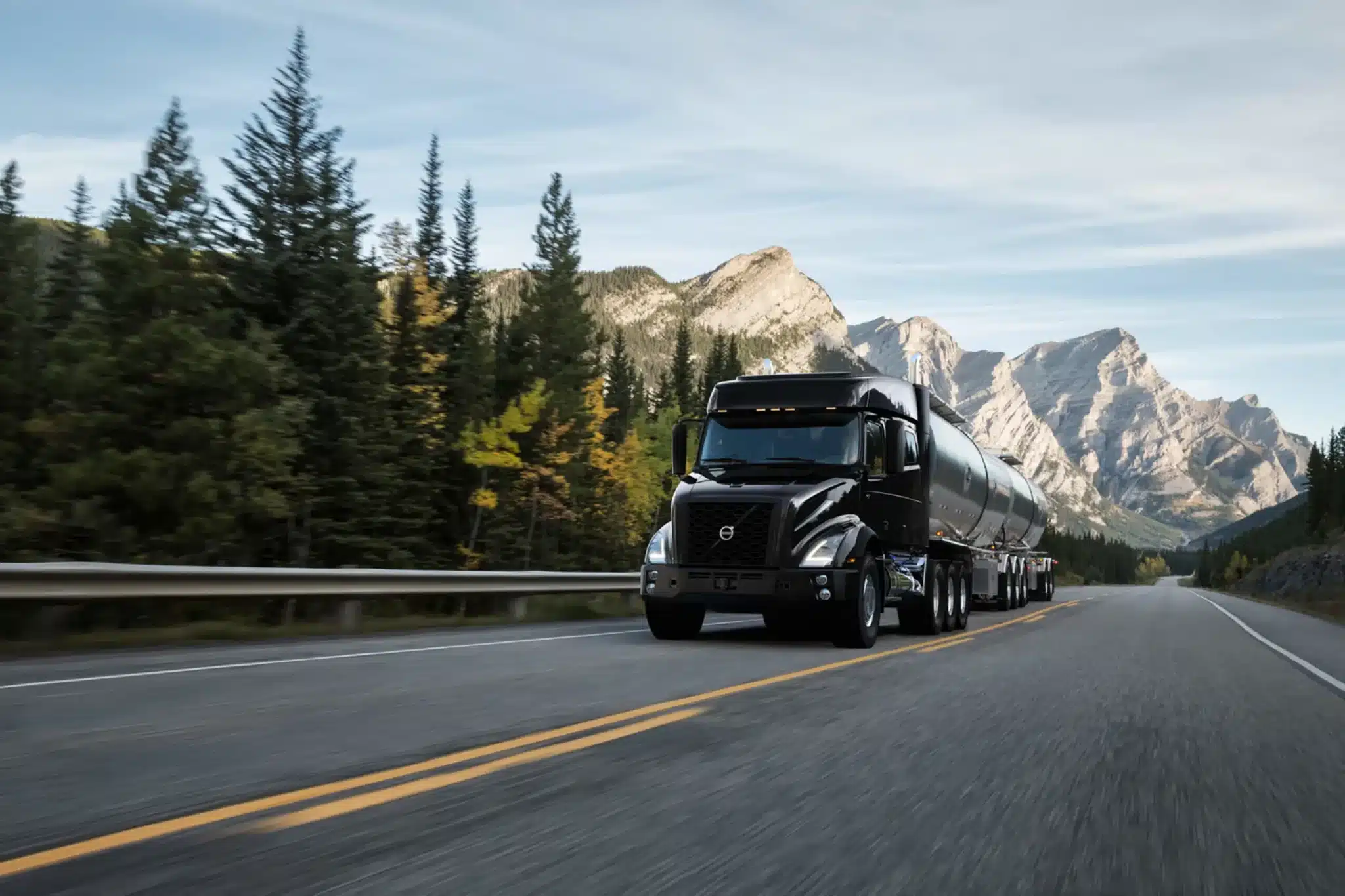 Black Volvo VNX driving up a highway with large mountains in the background as well a trees