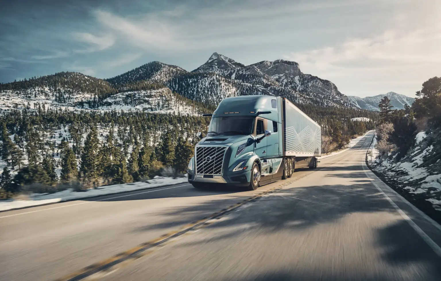 Blue all-new Volvo VNL 860 with a dry van driving up a mountain pass with trees and snow on the side of the road.