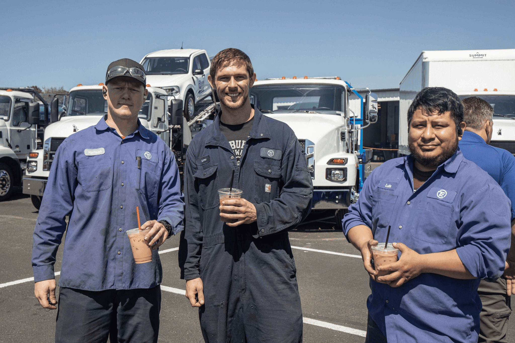 Three technicians smiling with coffee in their hands in front of trucks.