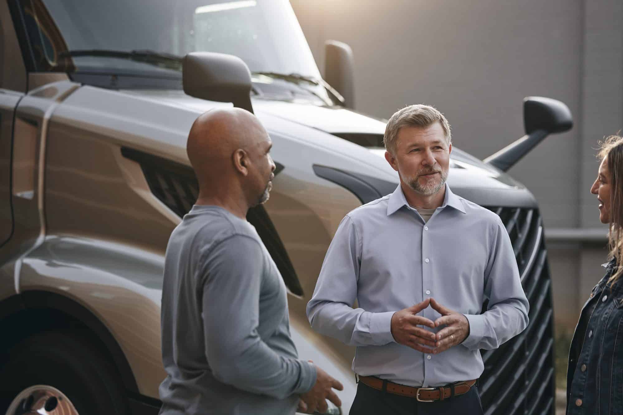 Two men speaking in front of a gold all-new VNL.