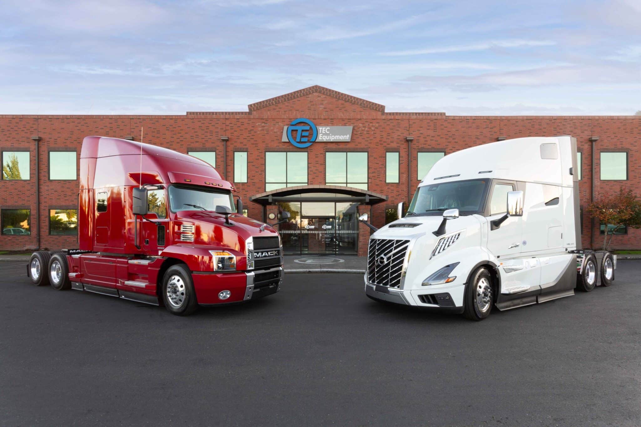 A red Mack Anthem and a white all-new Volvo VNL facing each other parked in front of TEC Equipment headquarters.